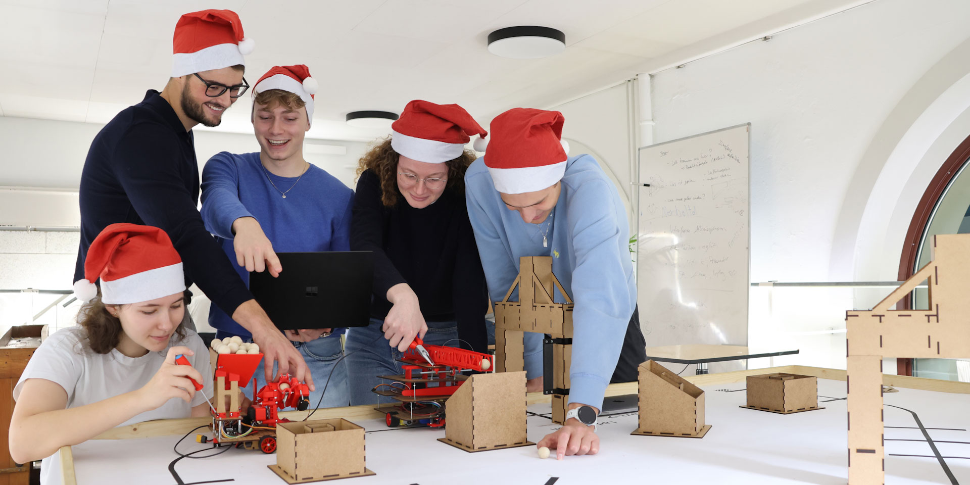 5 Studierende mit weihnachtlichen Zipfelmützen, die an kleinen Robotern basteln