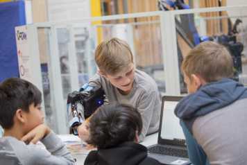 Vergrösserte Ansicht: Vier Kinder mit einem Roboter-Arm arbeiten am Laptop, um ihn zu programmieren