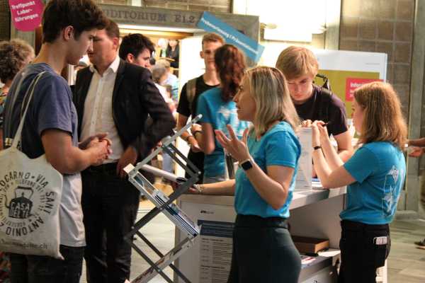 Maturand spricht mit einer Mitarbeiterin der Studienadministration am Infostand. Im Hintergrund sind noch viele andere Menschen zu sehen.