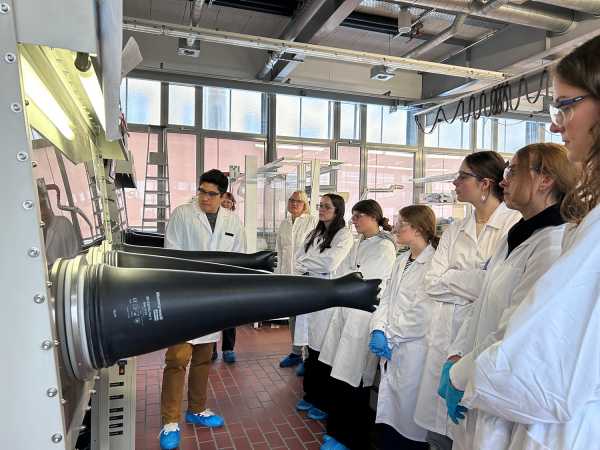 A doctoral student explains lab equipment, while young women pay close attention, everybody is wearing lab coats and protective glasses